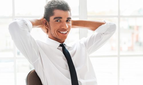 Make a small pause in your work! Handsome young Afro-American man in shirt and tie holding hands behind head and smiling while sitting at his working place