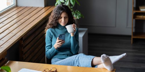Procrastination in daily working life. Young happy smiling female employee sitting at office desk with mobile phone, being distracted from work, relaxing and chatting online with friend during workday