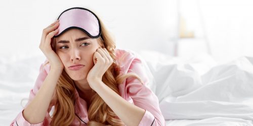 Upset tired bored lady wake up after bad sleep, tired or sleepless. Upset sad unhappy beautiful millennial european female in pink pajama and sleep mask lies on white bed in bedroom, empty space